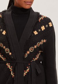Black knit cardigan with intricate floral embroidery in yellow, orange, and gray. Features a tie belt and frayed edges at the collar.