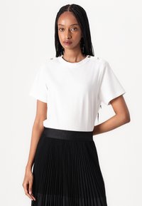 White t-shirt with short sleeves and metallic button details on shoulders, paired with a black pleated skirt featuring a branded waistband.