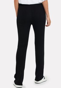 Zwarte geribbelde broek met een aansluitende taille, uitlopende pijpen en een gestructureerde stof. Gekleed met witte schoenen. Achteraanzicht dat de lengte en pasvorm toont.