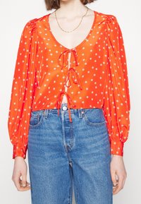 Femme portant un chemisier rouge à fleurs à manches longues avec trois liens à l'avant et un jean taille haute bleu, ainsi qu'un collier chaîne simple.