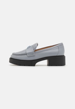 Mocasín de cuero gris claro con tacón y suela negros y gruesos, con una correa cosida sobre el empeine y diseño de puntera mocasín.