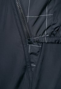 Tejido de chaqueta negra con una cremallera visible. El interior presenta un fruncido elástico y un patrón de rejilla cosido con hilo blanco. Acabado texturizado.