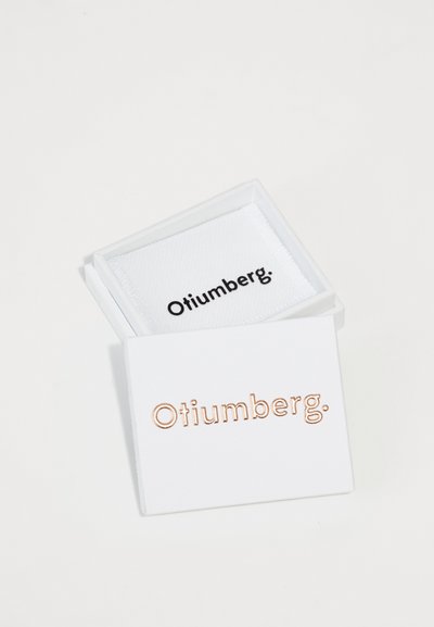 Otiumberg smykkeskrin, hvidt stof med roséguld bogstaver. Skrinet er lukket; logo i sort på en hvid baggrund indeni. Rektangulær form.