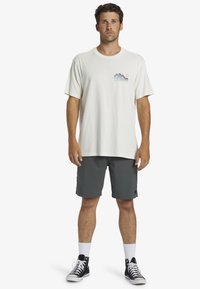 Men's light beige cotton t-shirt with mountain graphic, paired with dark gray shorts. Black high-top sneakers and white socks complete the look.
