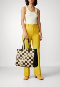 Stor vävd tote-väska med svarta detaljer, med ett upprepande logomönster och vit branding. Modellen bär gula högmidjade byxor.