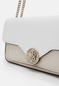 Sac à main en cuir blanc avec une bandoulière en chaîne dorée et un fermoir circulaire en or gravé de "GUESS 1981 LOS ANGELES".