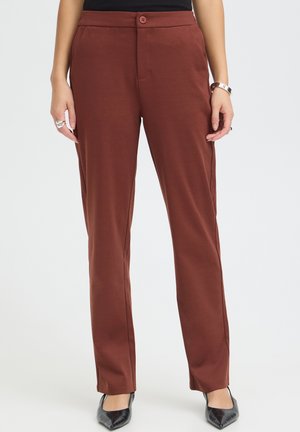 Femme portant un pantalon marron droit avec une fermeture par bouton et zip, ainsi que des chaussures noires à bout pointu, debout devant un fond uni.