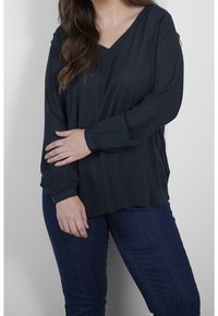 Blouse manches longues bleu foncé avec un décolleté en V et un tissu texturé. Présente une coupe décontractée et des poignets froncés. Associée à un jean foncé.