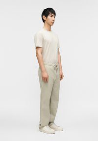 Ανοιχτό μπεζ κοντομάνικο μπλουζάκι με διακριτή υφή, σε συνδυασμό με ανοιχτό γκρι παντελόνι με ελαστική μέση και κρεμ αθλητικά παπούτσια, φορεμένα από έναν όρθιο άνδρα.