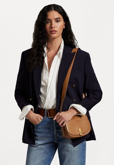 Pruunist nahast ristkeha kott, millel on kõver kuju ja kuldsetestdetailidest aksessuaarid, kantud tumesinise bleiseri ja valge särgi peal, kombineeritud siniste teksadega.