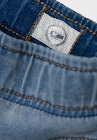 Denimstoff med blå farge og en falmet tekstur. Det er en knapp med en gjennomsiktig deksel og kontrasterende brune sømmer.