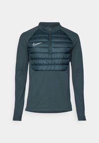 Mørkegrønn genser med lange ermer og glidelås i halsen, med en quilted puffer front, Nike-logo på venstre bryst, og glatte stofferm. 