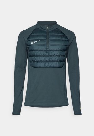 Pullover escuro de manga longa verde com gola em zíper, apresentando um painel frontal acolchoado, logótipo da Nike no lado esquerdo do peito e mangas de tecido suave.