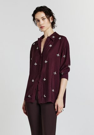 Donna con i capelli corti e scuri che indossa una blusa viola intenso con decorazioni di api argento e pantaloni scuri coordinati, su uno sfondo neutro.