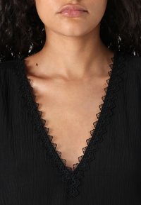 Black fabric top featuring a deep V-neck with intricate black lace trim. The texture is slightly crinkled, enhancing the design details.