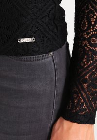 Schwarzes Spitzentop mit kunstvollen Mustern und einem silbernen Logo-Akzent, kombiniert mit dunkelgrauen Jeans, die sichtbare Nähte und ein Knopfdetail aufweisen.