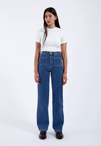 Jeans blu a vita alta con design a gamba dritta, caratterizzati da due grandi tasche anteriori e cuciture a contrasto, abbinati a una maglietta bianca aderente.