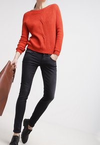 Suéter rojo de punto con costillas y escote redondo, combinado con jeans negros ajustados y zapatos negros con cordones. Incluye bolso de cuero marrón.