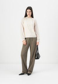 Kvinna iklädd en krämfärgad blus i spets med lång ärm, olivgröna vida byxor, svarta spetsiga flats, som håller en svart handväska.