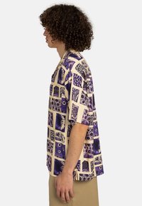 Short-sleeve shirt in beige fabric featuring a purple geometric pattern with various shapes and textures, displayed from a side perspective.
