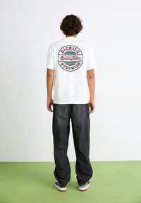 Hvit bomull T-skjorte med et sirkulært grafikkdesign på baksiden, som viser en lastebil og teksten "DICKIES WORKWEAR", kombinert med løse svarte bukser.