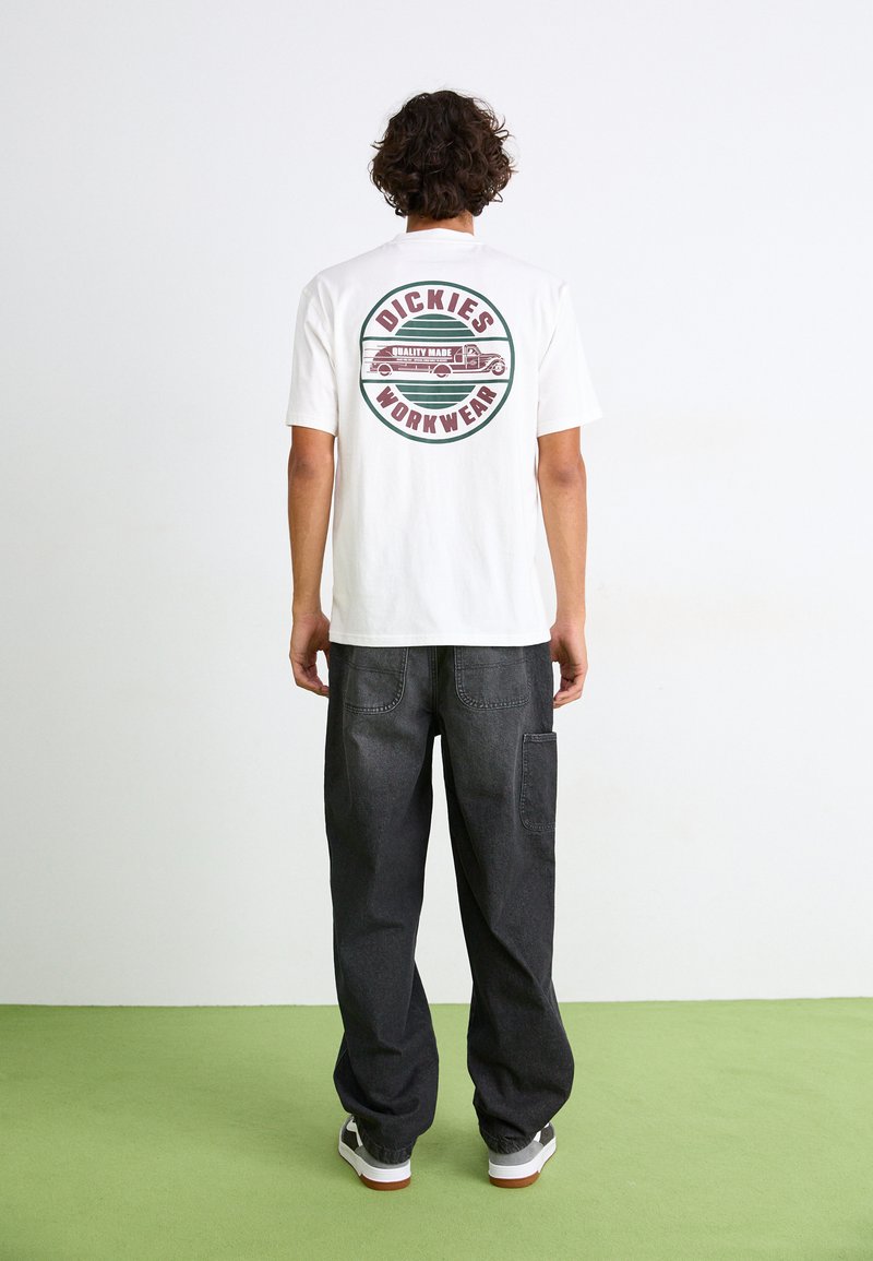 Weißes Baumwoll-T-Shirt mit einem runden Grafikdesign auf dem Rücken, das einen Truck und den Text "DICKIES WORKWEAR" zeigt, kombiniert mit lockeren schwarzen Hosen.