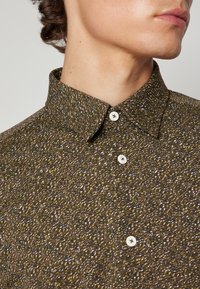 Camicia marrone con un motivo testurizzato che presenta piccole macchie bianche e gialle. Include un colletto rigido e chiusure a bottone bianche.