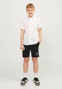 Vit kortärmad skjorta med knappar, svarta shorts med logodetalj, vita strumpor och svarta sneakers med beige detaljer. Avslappnad outfit för barn.