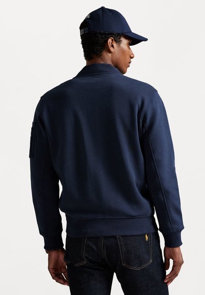 Marineblauwe sweatshirt met een geribde kraag en manchetten, voorzien van een zijzak op de arm en een gladde textuur, gecombineerd met donkere spijkerbroeken.