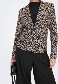 Femme portant un blazer imprimé léopard sur un top noir et un pantalon noir à jambes larges, tenant un sac à main noir avec une sangle tressée.