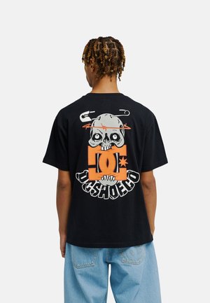 Osoba ubrana w czarną koszulkę z dużą grafiką czaszki, agrafką, pomarańczowym logo i napisem DC SHOES CO na plecach, połączoną z niebieskimi dżinsami.