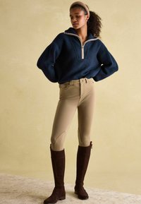 Marineblauer Pullover mit einem halben Reißverschluss und strukturiertem Finish, kombiniert mit hellbeigen, enganliegenden Reithosen und braunen, kniehohen Wildlederstiefeln.