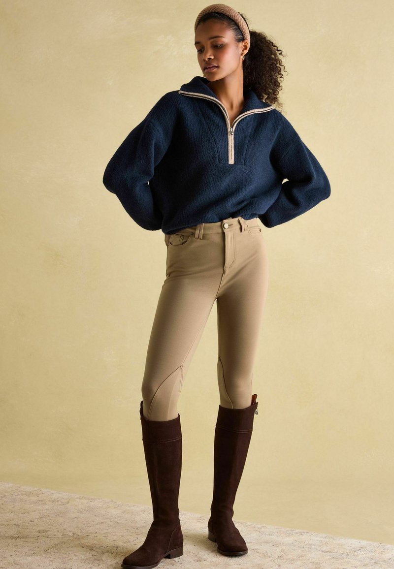 Marineblauer Pullover mit einem halben Reißverschluss und strukturiertem Finish, kombiniert mit hellbeigen, enganliegenden Reithosen und braunen, kniehohen Wildlederstiefeln.