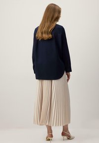 Femme aux cheveux longs portant une chemise bleu marine à manches longues, une jupe midi plissée beige et des escarpins slingback beiges, debout devant un fond uni.