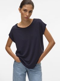 Mladá žena s tmavými vlasy sepnutými do drdolu, oblečená v volném krátkém modrém tričku s krátkým rukávem a světlých modrých džínech, s rukama v kapsách.