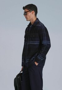 Veste chemise à carreaux noir et bleu avec deux poches poitrine et un bouton de fermeture, portée sur un haut noir, associée à un pantalon marine et un sac noir.