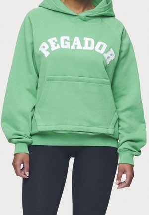 Personne portant un sweat à capuche vert avec le mot « PEGADOR » en lettres blanches, une poche kangourou à l'avant et un legging noir.