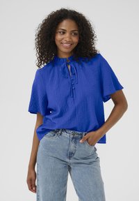 Blouse bleue à texture, à manches courtes, avec un détail à nouer au cou et une fermeture à boutons. Associée à un jean droit en denim délavé.