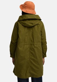 Olivgrön, lätt parka jacka med huva, indragen midja och långa ärmar. Accentuerad med en texturerad orange bucket hat.