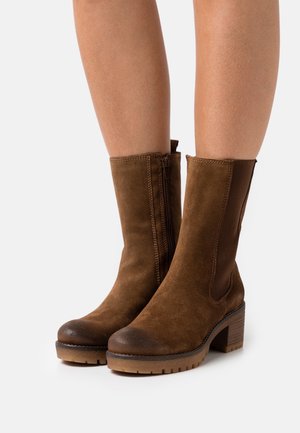 Bottines en suède marron avec un talon empilé, des panneaux latéraux élastiques et une fermeture éclair sur le côté. Finition texturée remarquable et coutures contrastantes.