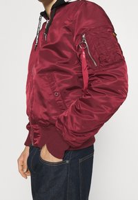 Giacca bomber color bordeaux con finitura satinata lucida, polsini a coste e una tasca con zip sulla manica. Presenta un gancio per appendere.