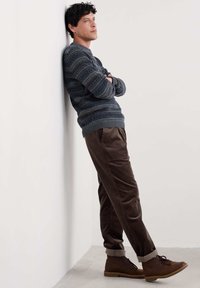 Vīrietis ar rakstainu džemperi, brūniem kordūrveida biksēm un brūnām zābakiem atspiedies pret baltu sienu ar saliktām rokām, domīgi raugās augšup.