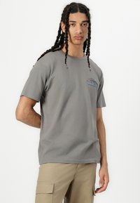 Graues Baumwoll-T-Shirt mit kurzen Ärmeln, mit einem blauen und braunen grafischen Logo auf der linken Brust. Das Modell trägt beige Cargo-Shorts.