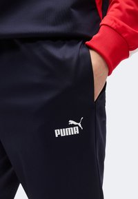 Άτομο που φοράει μαύρο παντελόνι Puma με το χέρι στην τσέπη και εμφανές κόκκινο μανίκι πάνω σε ανοιχτό φόντο.