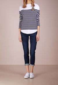 Persona che indossa una maglietta a righe navy e bianche a maniche lunghe, jeans scuri arrotolati e sneakers bianche, in piedi contro uno sfondo neutro.