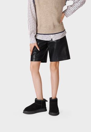 Persona che indossa pantaloncini in pelle neri, stivaletti neri, camicia a righe e maglione beige senza maniche, in piedi contro uno sfondo semplice.