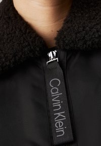 Giacca nera con colletto peloso, dotata di zip e di un'etichetta intrecciata con "Calvin Klein" in testo grigio sulla parte anteriore.
