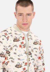 Floral bomberjack met een crèmekleurige basis en levendige rode en groene tropische patronen, ritssluiting aan de voorkant en gebreide groene kraag.