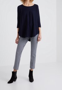 Frau trägt ein lockeres navy-blaues Oberteil, schwarze und weiße karierte 3/4-Hosen und schwarze Wildleder-Stiefeletten und steht auf einem schlichten weißen Boden.