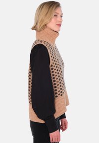 Beige strikket vest med svarte geometriske mønstre, høy ribbestrikket krage og svart langermet skjorte under.